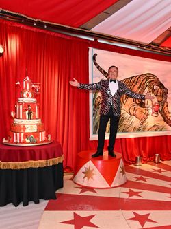 Man in striped jacket stands on circus platform with circus cake and tiger mural against red curtains at event space.