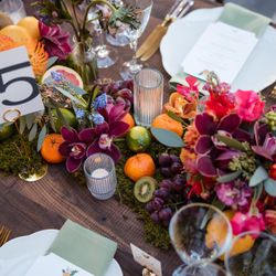 Table setting with floral and fruit centerpiece, candles, menus, and green cloth napkins on wooden surface
